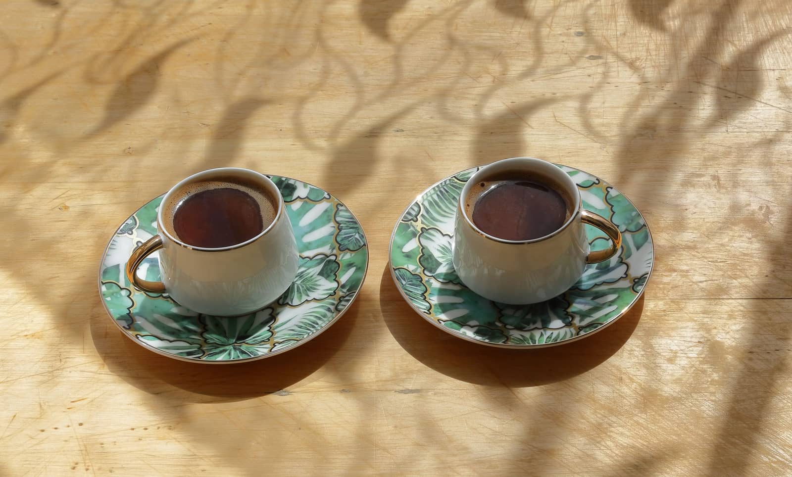 Two patterned coffee cups and saucers on a wooden table, dappled leaf shadows falling across the wood.