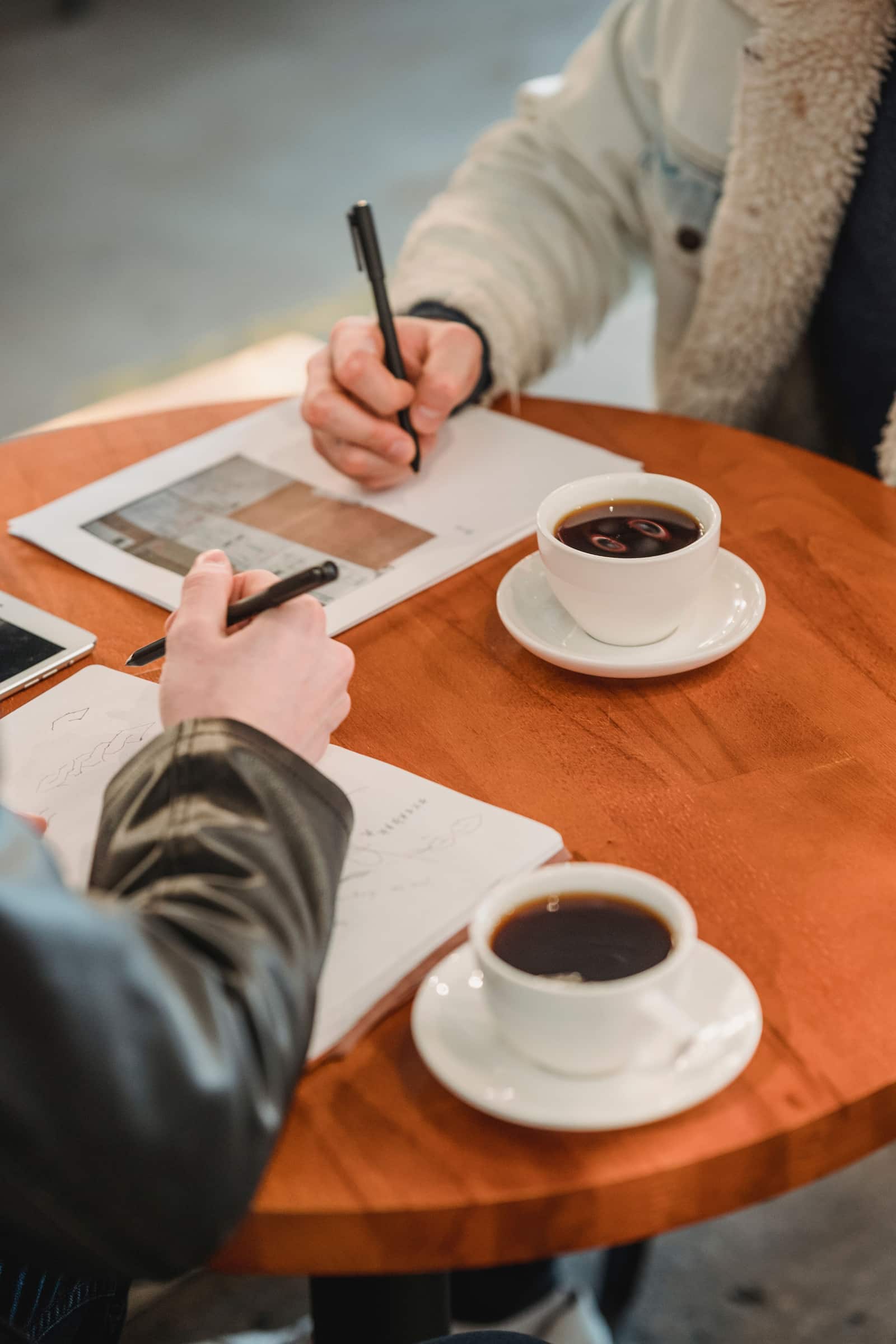Two people at a cafe table with notebooks, sketches and two cups of coffee, hands visible but faces out of frame.