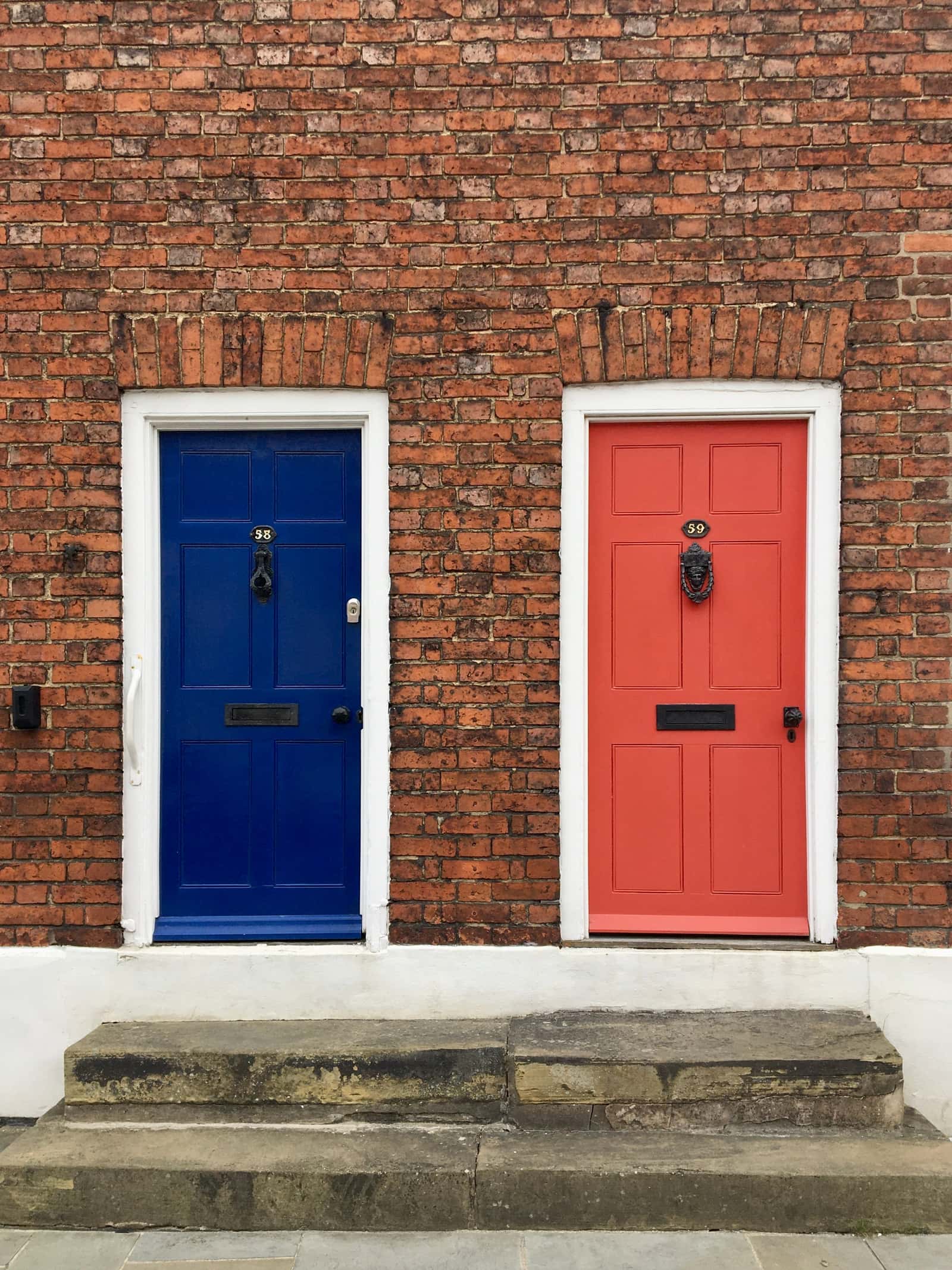 Two front doors side by side in a brick terrace, one navy blue and one coral, each with its own house number.