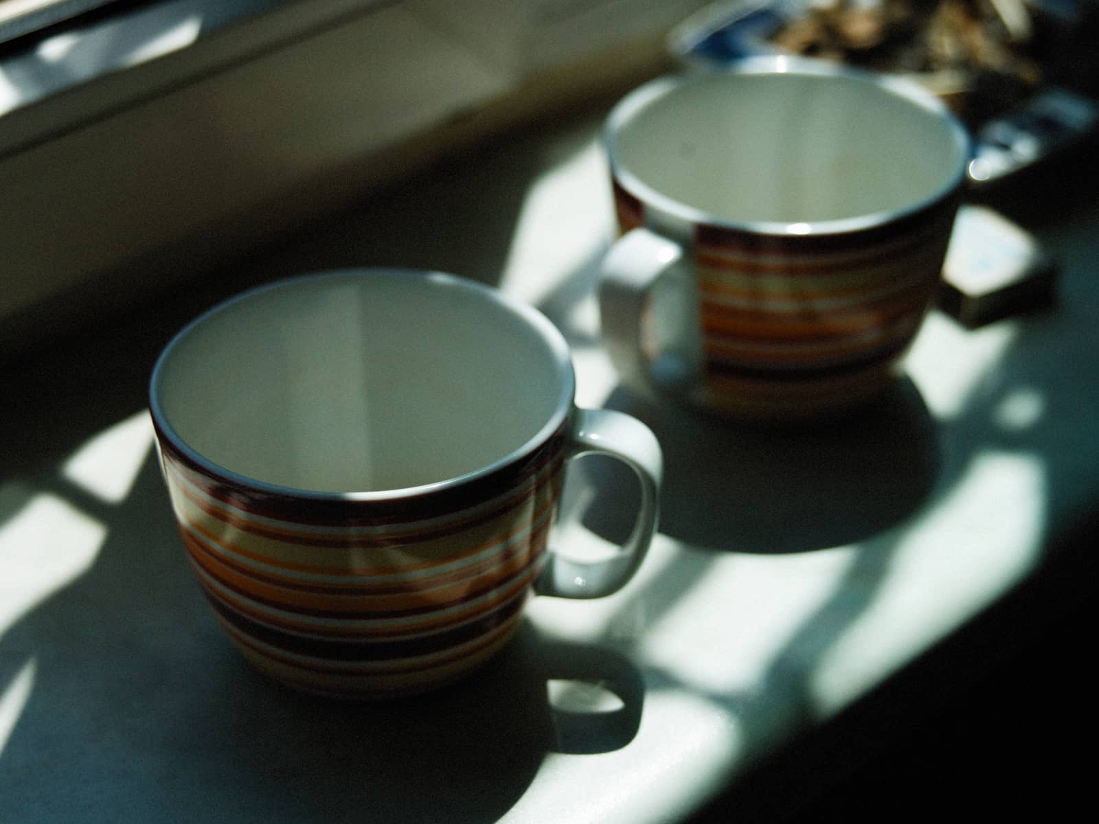 Two striped mugs on a sunlit windowsill, morning light casting long shadows across the sill.